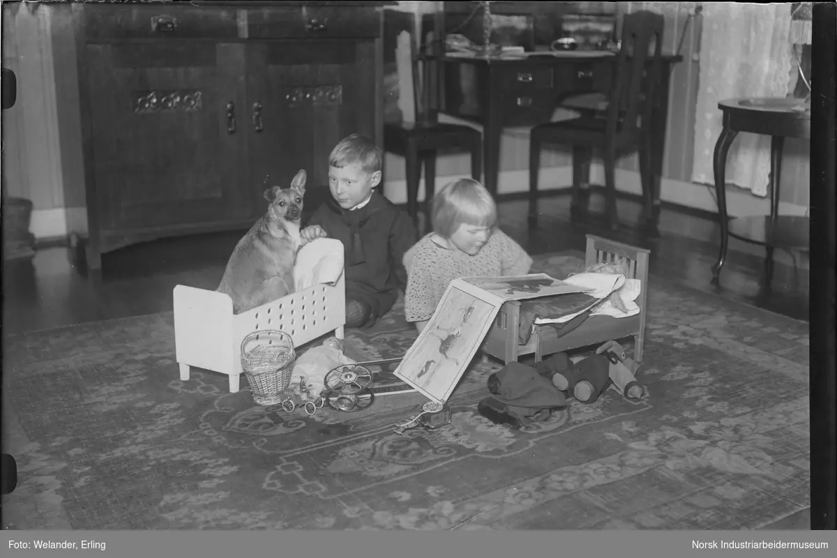 To barn leker på gulv i stue. Jenta sitter med en billedbok liggende over dukkeseng. Gutten har en hund i en dukkeseng. Leker liggende på gulvet rundt dukkesengene. Stuemøblement i bakgrunnen.
