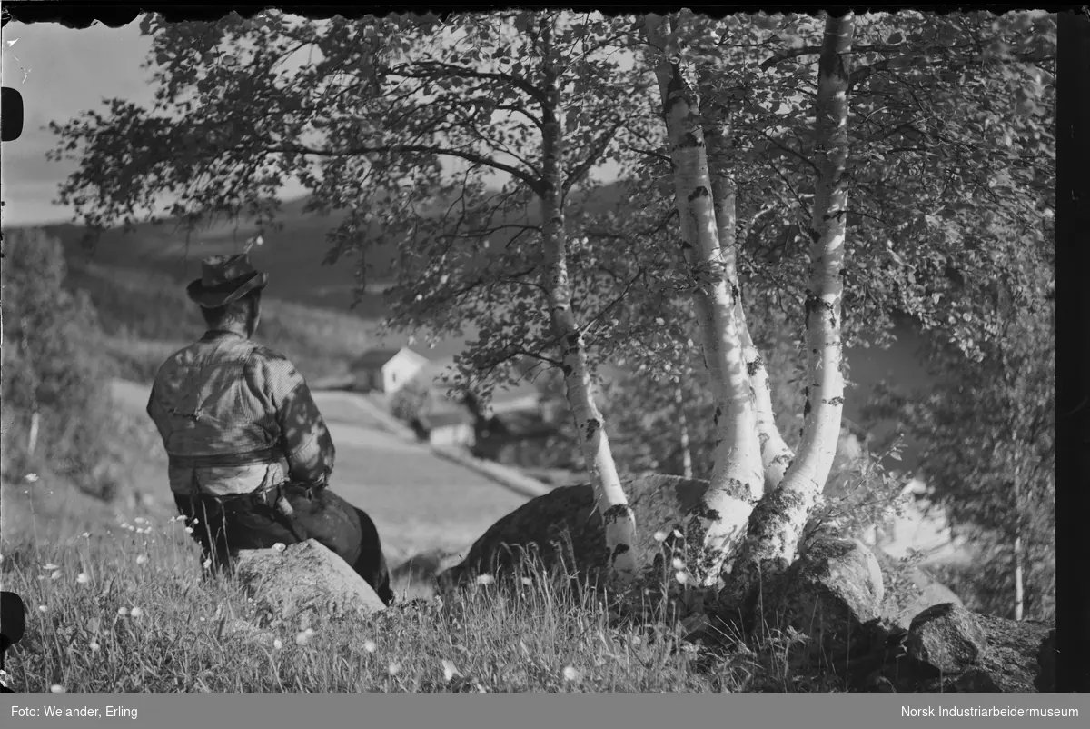 Mann sittende på en høyde og se utover bygninger. Mannen har hatt, skjorte og vest og kniv i belte på buksa.