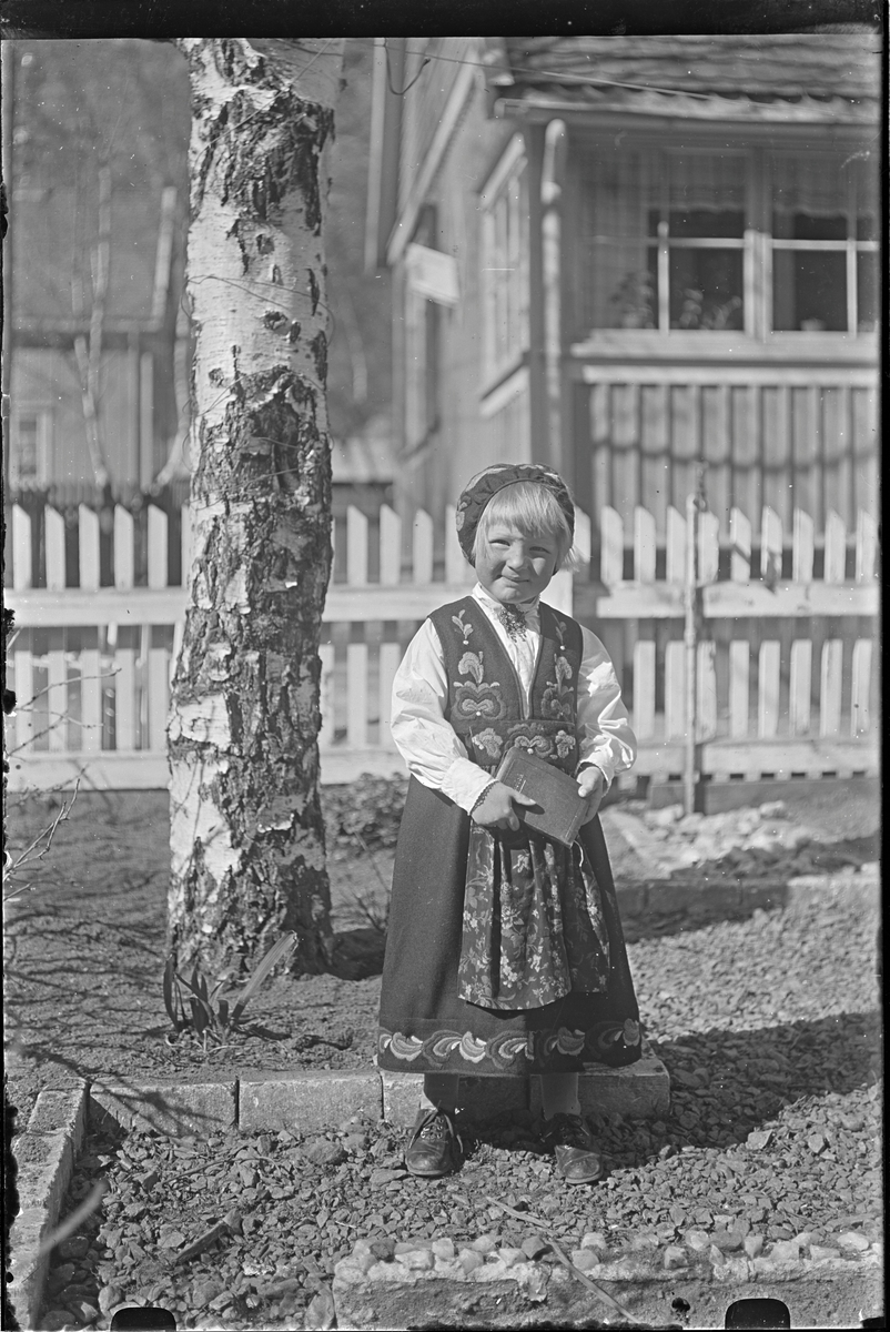 Jente stående ved tre kledd i bunad med kyse på hodet. Jenta har en bok i hendene. Bolighus med stakittgjerde rundt i bakgrunnen.