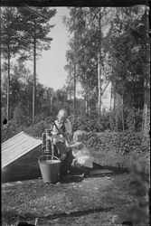 Jente og gutt utendørs ved brønn. De pumper vann opp i en bø