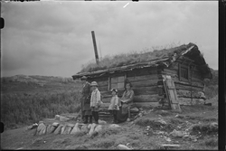 To kvinner og to barn stående utenfor laftet hytte kalt "Hyg