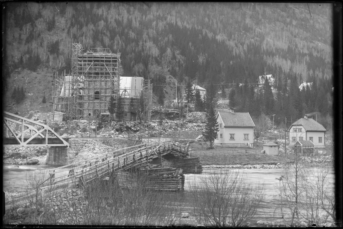 Rjukan kirke under bygging i 1915. Trestilas rundt kirken, brakker stående på forsiden av kirkebygget. Avstivning for tørrmuring av buet vindu ligger ved siden av brakke. Bebyggelse i Sam Eydesgate (Blindtarmen) og Skogveien i bakgrunnen. Trebro over Måna til høyre for jernbanebroen Mæland bro.