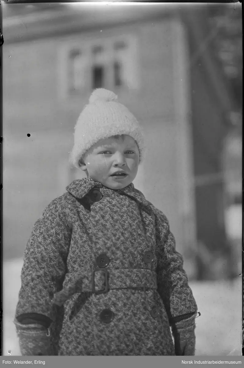 Utendørs portrett. Barn kledd i vinterklær stående på utsiden av bolighus.
