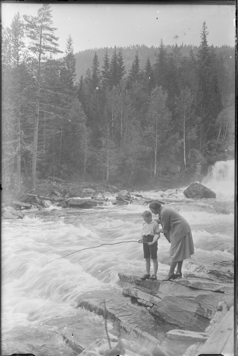 Kvinne og barn stående ved elvestryk. Gutten holder en hjemmelaget fiskestang i hendene. Kvinnen er kledd i kjole og høye hæler, gutten i shorts og skjorte.