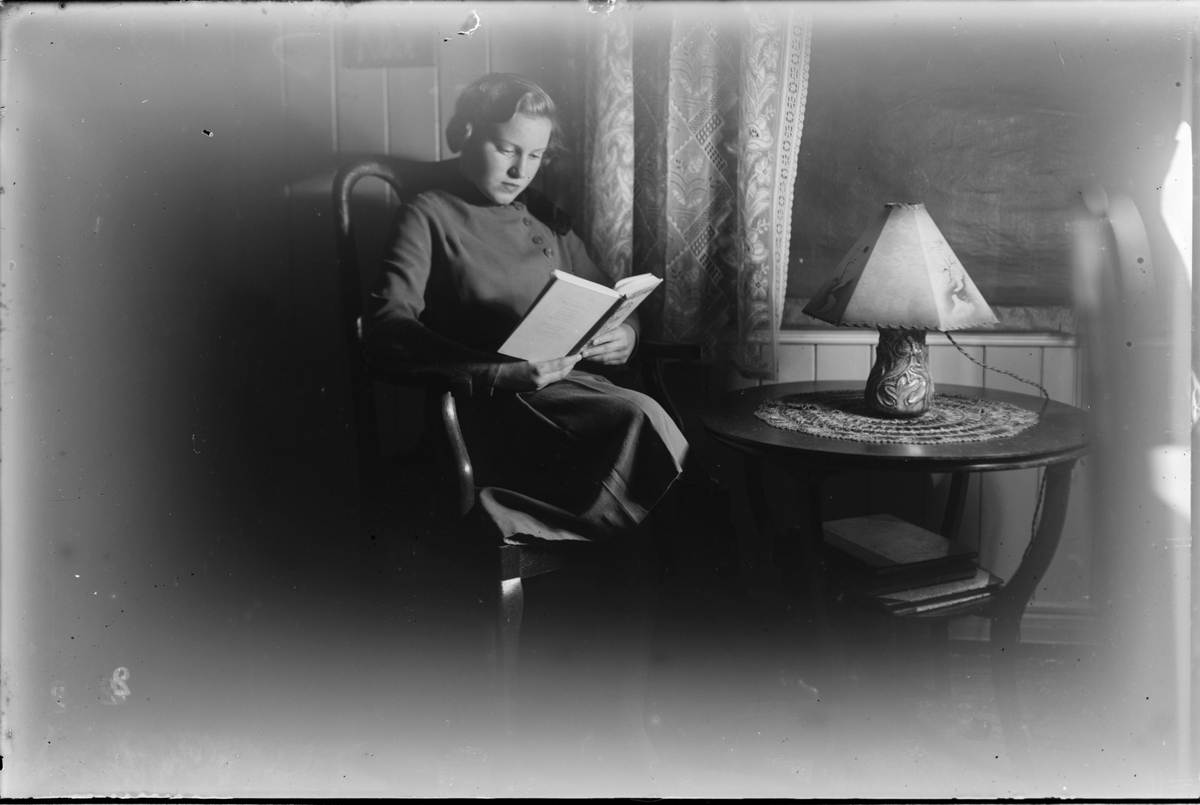Kvinne sittende i stue og lese bok. Heklede gardiner og blendingsgardin foran vinduet. Rundt bord med lampe på foran vinduet.