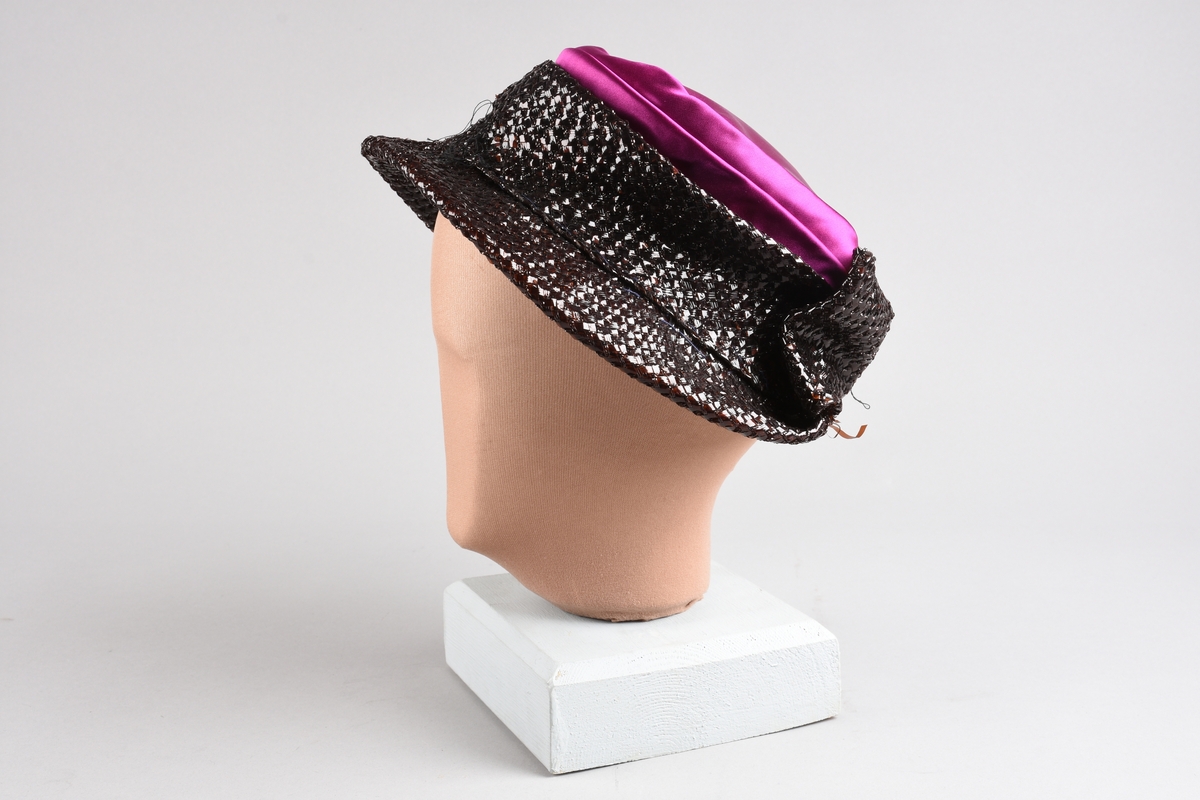 En purpurfarget hatt av flat satengpull med folde-drapert kant, omspennes av en lav, kobbersort, flettet cellofan-"karm" og smal brem. I nakken er det en bred, flat sløyfe av cellofanstoffet. Bremmen er senere avskåret i nakken og sydd opp. Over pannen viser trådender at noe pynt er tatt av (rosene og strutsefjæren på den senere hatt GB 18344?). Inni er hatten avstivet med stramei-liknende stivstoff og er isydd et hattebånd.