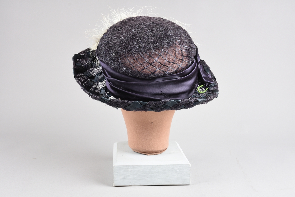 Mørkfiolett, flatpullet og bredbremmet hatt i  "bretonne-" el. "canotier-"fasong. Laget av nettvevede, kunstige stråbånd. Rundt pullen er et "hattebånd" av bred drapert sateng i samme farge, festet med spenne av hamret messingblikk. Senere laget om til kanutt-kostyme, med kunstig rosegren oppå offwhite strutsefjær dandert over pannen. Inni pullen er en skulderpute satt fast med sikkerhetsnål.  Hårnål og hatte-elastikk til feste av hatten.