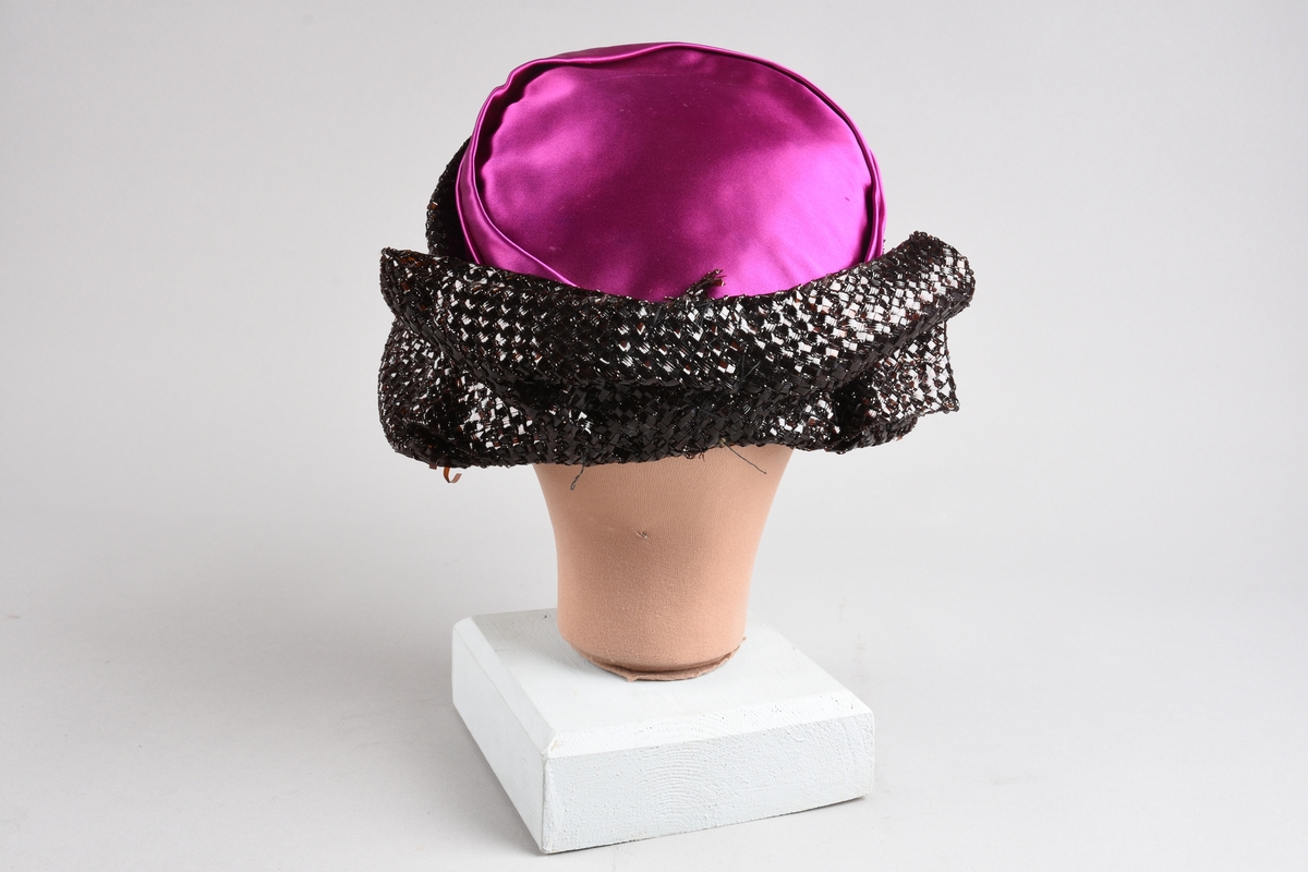 En purpurfarget hatt av flat satengpull med folde-drapert kant, omspennes av en lav, kobbersort, flettet cellofan-"karm" og smal brem. I nakken er det en bred, flat sløyfe av cellofanstoffet. Bremmen er senere avskåret i nakken og sydd opp. Over pannen viser trådender at noe pynt er tatt av (rosene og strutsefjæren på den senere hatt GB 18344?). Inni er hatten avstivet med stramei-liknende stivstoff og er isydd et hattebånd.