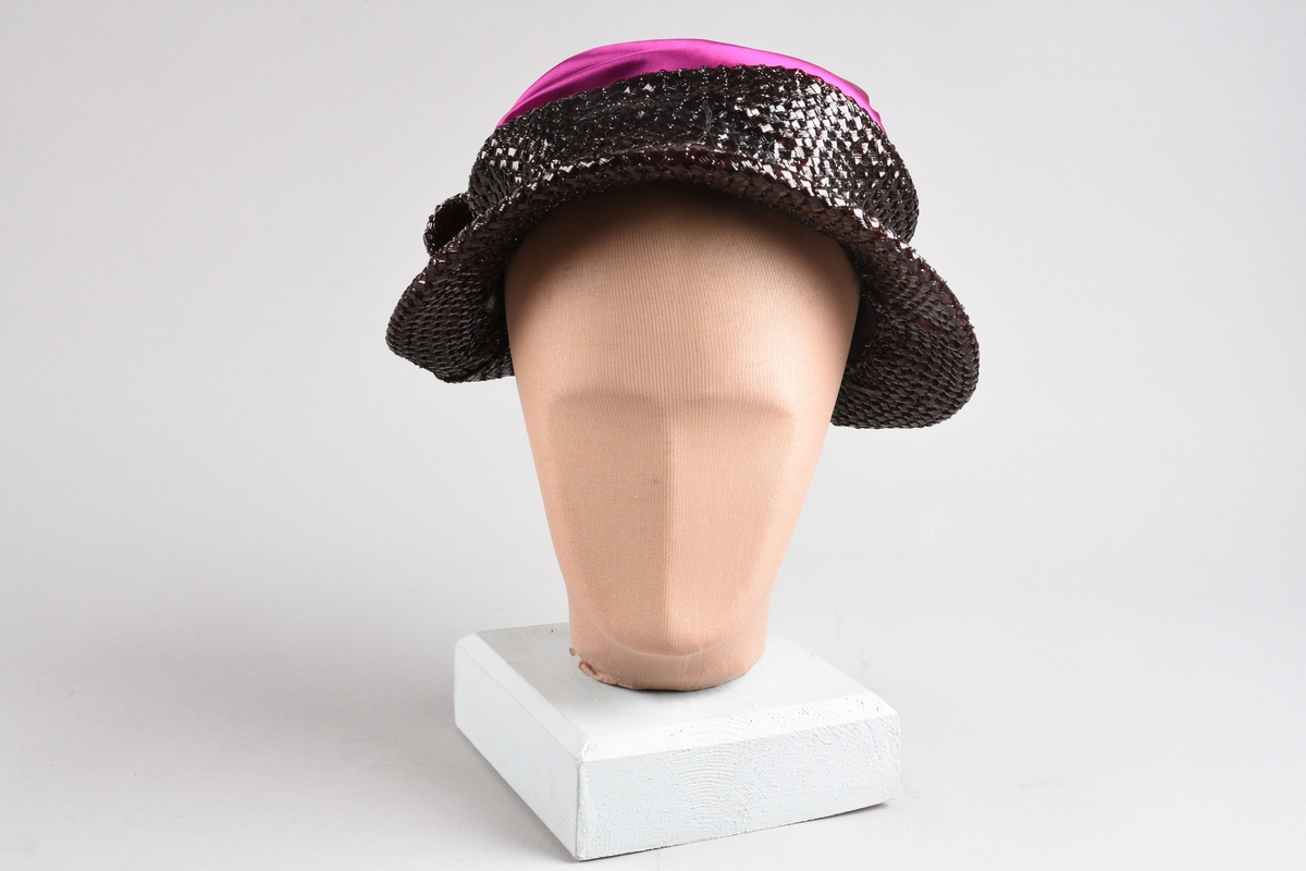 En purpurfarget hatt av flat satengpull med folde-drapert kant, omspennes av en lav, kobbersort, flettet cellofan-"karm" og smal brem. I nakken er det en bred, flat sløyfe av cellofanstoffet. Bremmen er senere avskåret i nakken og sydd opp. Over pannen viser trådender at noe pynt er tatt av (rosene og strutsefjæren på den senere hatt GB 18344?). Inni er hatten avstivet med stramei-liknende stivstoff og er isydd et hattebånd.