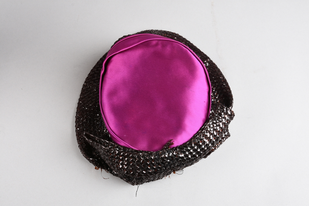 En purpurfarget hatt av flat satengpull med folde-drapert kant, omspennes av en lav, kobbersort, flettet cellofan-"karm" og smal brem. I nakken er det en bred, flat sløyfe av cellofanstoffet. Bremmen er senere avskåret i nakken og sydd opp. Over pannen viser trådender at noe pynt er tatt av (rosene og strutsefjæren på den senere hatt GB 18344?). Inni er hatten avstivet med stramei-liknende stivstoff og er isydd et hattebånd.