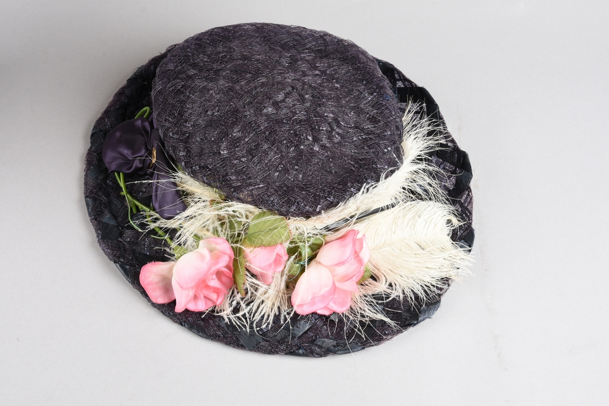 Mørkfiolett, flatpullet og bredbremmet hatt i  "bretonne-" el. "canotier-"fasong. Laget av nettvevede, kunstige stråbånd. Rundt pullen er et "hattebånd" av bred drapert sateng i samme farge, festet med spenne av hamret messingblikk. Senere laget om til kanutt-kostyme, med kunstig rosegren oppå offwhite strutsefjær dandert over pannen. Inni pullen er en skulderpute satt fast med sikkerhetsnål.  Hårnål og hatte-elastikk til feste av hatten.