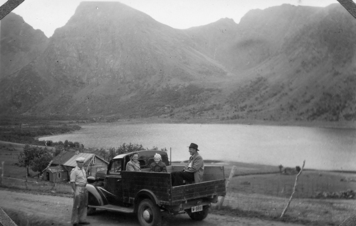 Fra Slåttnes i Hadsel med Slåttnesbogen i bakgrunnen. På nedsiden av veien i bakgrunnen huset til Andor og Helga Mathisen. I kassen på latebilen, som er en ca 1937 Chevrolet ombygd til lastebil, sitter Aslaug og Ingvald Johnsen fra Fleines og deres datter Audhild. På siden av bilen står Heggelund Bjørhovde fra Bø. Bildet er fra første halvdel av 1950-tallet.