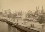 Sundsvalls stadsbrand 1888. Kvar efter brandens framfart finns bara skorstenspipor och utbrända huskroppar. I bildens framkant syns Selångersån och bland husresterna står några män varav två i uniform.