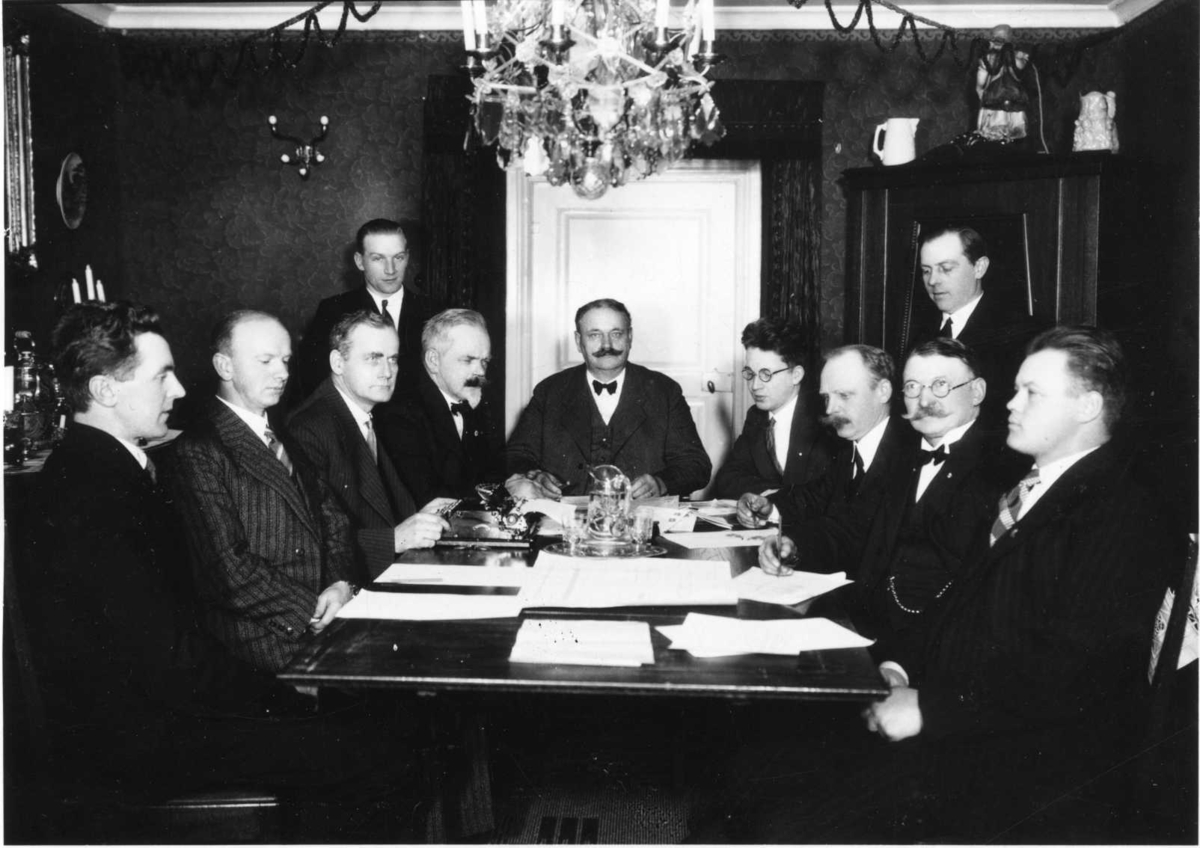 Nio män sitter runt ett bord med papper framför sig, två män står intill.