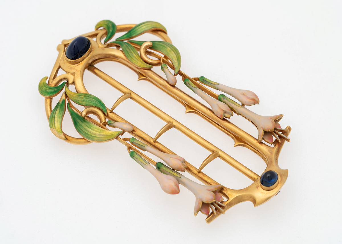 Ovalformet gjennombrutt spenne i gull, dekorert med blåfargede steiner. Langs spennes langsider slynger det seg grønnfargede blader samt jasminer i hvit og blekrosa emalje.
