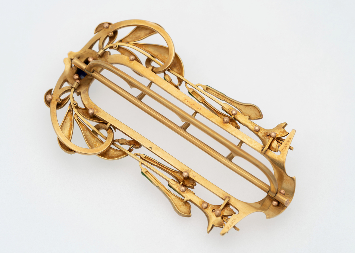 Ovalformet gjennombrutt spenne i gull, dekorert med blåfargede steiner. Langs spennes langsider slynger det seg grønnfargede blader samt jasminer i hvit og blekrosa emalje.