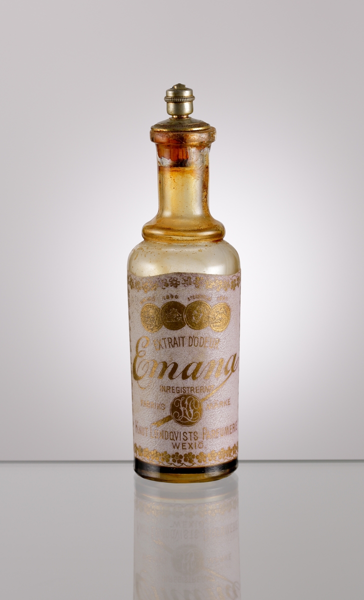Parfymflaska från Knut Lundqvists Kemisk-tekniska fabrik i Växjö (se bifogat foto) med etikett och metallförsedd kork. Text på etikett: "EXTRAIT D'ODEUR // Emana // INREGISTRERAT FABRIKS MÄRKE KL // KNUT LUNDQVIST PARFUMERI // WEXIÖ".