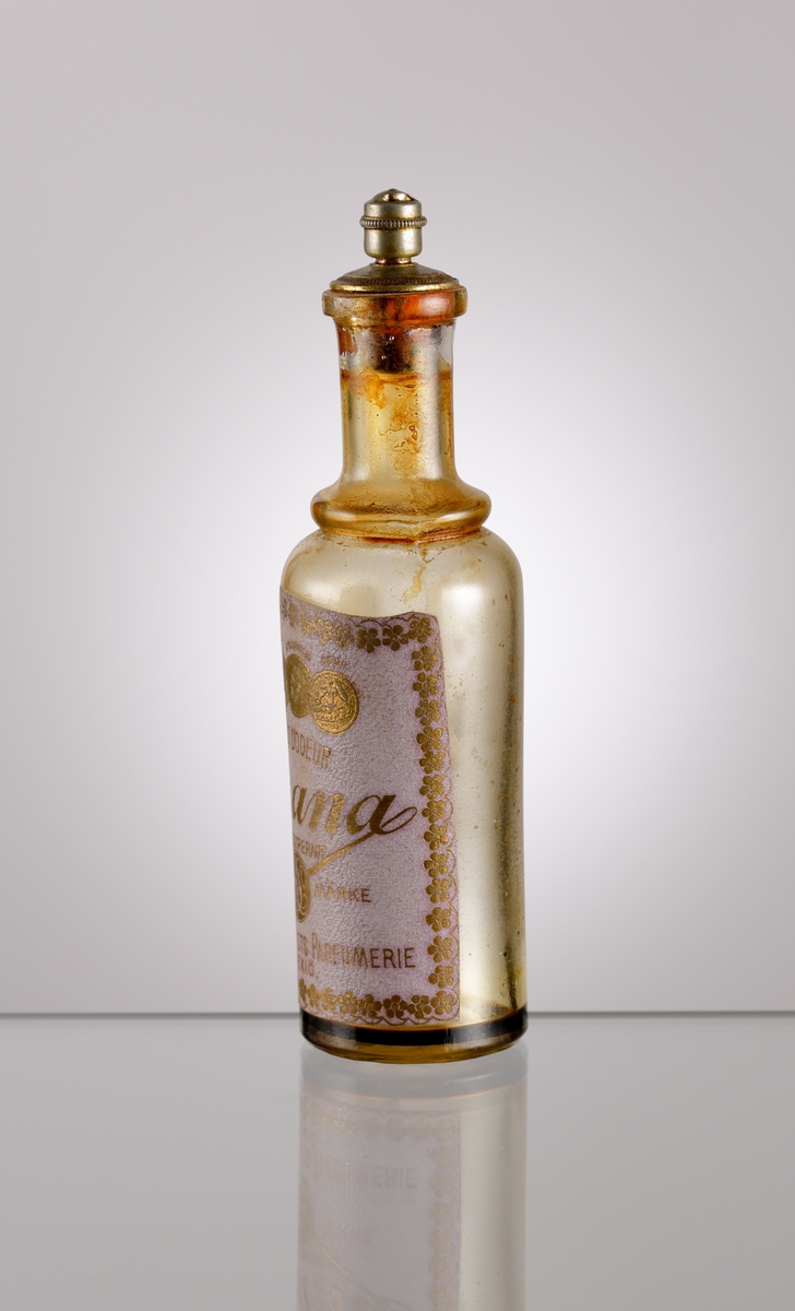 Parfymflaska från Knut Lundqvists Kemisk-tekniska fabrik i Växjö (se bifogat foto) med etikett och metallförsedd kork. Text på etikett: "EXTRAIT D'ODEUR // Emana // INREGISTRERAT FABRIKS MÄRKE KL // KNUT LUNDQVIST PARFUMERI // WEXIÖ".