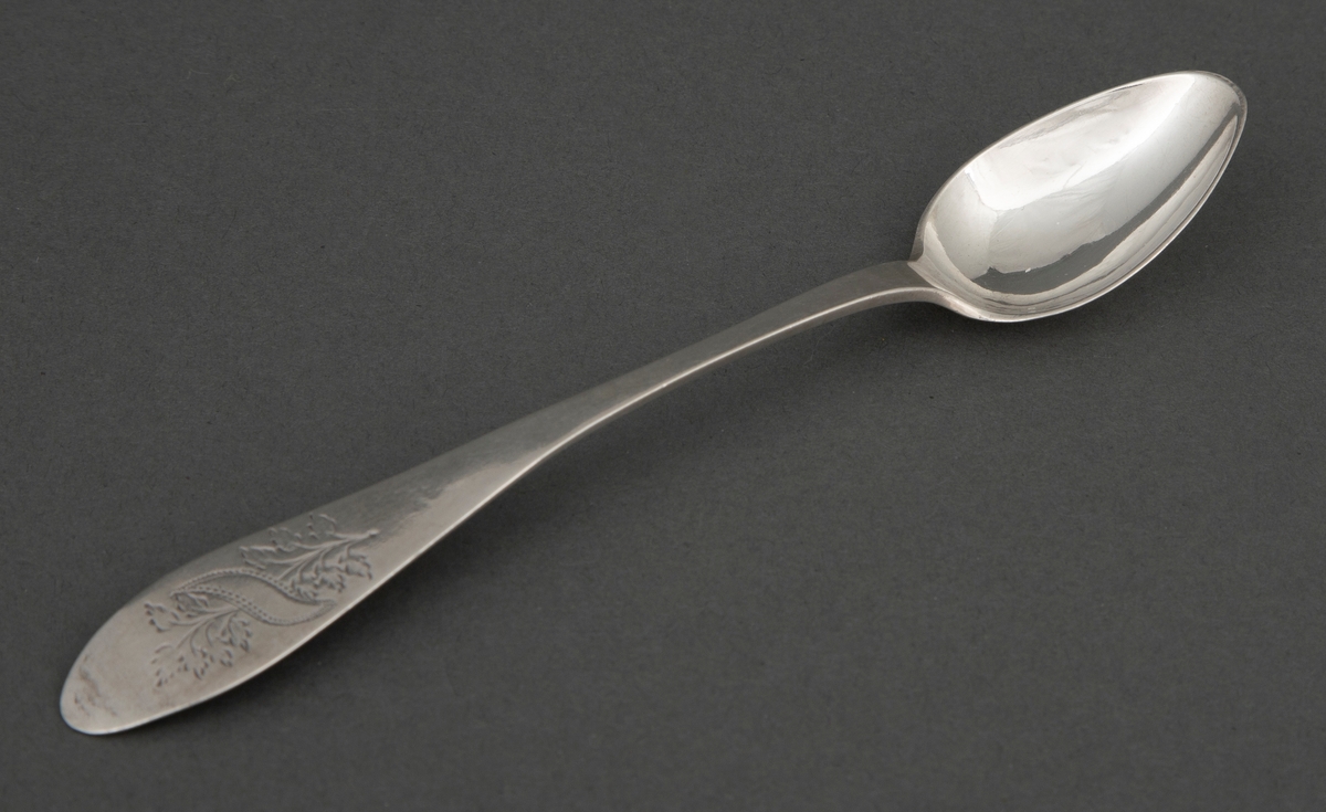 Skje i sølv. Lite dråpeformet blad. Skaftet er flatt med økende bredde mot skaftenden. Bredden tiltar mot den spisse, men avrundede skaftenden. I skaftenden finnes et gravert løvmotiv med skrått inskripsjonsfelt over.
