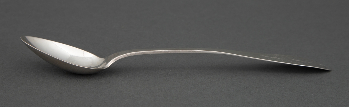 Skje i sølv. Lite dråpeformet blad. Skaftet er flatt med økende bredde mot skaftenden. Bredden tiltar mot den spisse, men avrundede skaftenden. I skaftenden finnes et gravert løvmotiv med skrått inskripsjonsfelt over.
