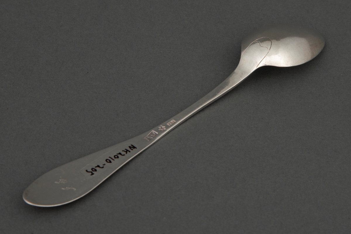 Skje i sølv. Lite dråpeformet blad. Skaftet er flatt med økende bredde mot skaftenden. Bredden tiltar mot den spisse, men avrundede skaftenden. I skaftenden finnes et gravert løvmotiv med skrått inskripsjonsfelt over.
