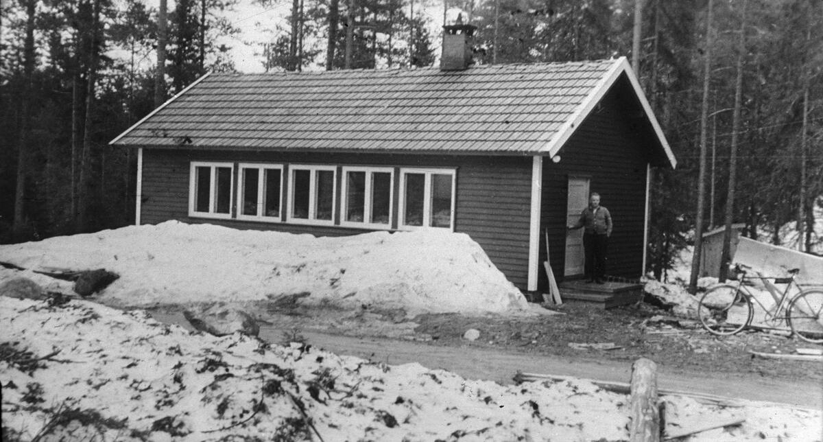 Redskapssentral hvor spesialister steller redskapen til et stort antall skogsarbeidere.  Enetasjes bygning i bindingsverk med horisontal panel og med sementsteintekket tak.  På langveggen er det fem tofags funkisvinduer.  Inngangsdøra er plassert på gavlveggen, hvor det står en mann på trappa.  Til høyre for inngangen står en sykkel.  Foran huset går det en veg.  I bakgrunnen står skogen tett. 

Dette fotografiet er fra en lysbildeserie Landbrukets film- og bildekontor produserte, antakelig i åra like etter 2. verdenskrig.  Med bildene fulgte det et lite stensilert teksthefte.  Her har dette fotografiet fått følgende tekst:

«Redskapssentral.  Her driver spesialister og steller redskaper for 100 arbeidere.»