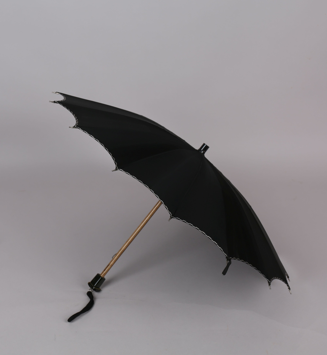 Paraply med 16 metallspilar. Stong i tre. Trekt med stoff i kunstfiber, ensfarga svart med dekor i sølv/svart rutemønster. Handtak i plast med hempe i tekstil til å ta inn på handa. Hempe for lukking av paraplyen.