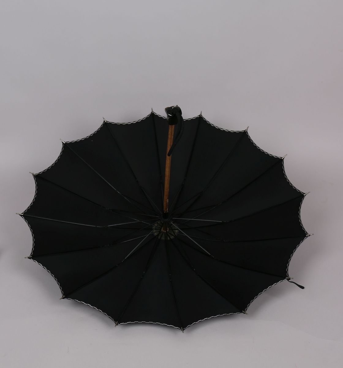 Paraply med 16 metallspilar. Stong i tre. Trekt med stoff i kunstfiber, ensfarga svart med dekor i sølv/svart rutemønster. Handtak i plast med hempe i tekstil til å ta inn på handa. Hempe for lukking av paraplyen.