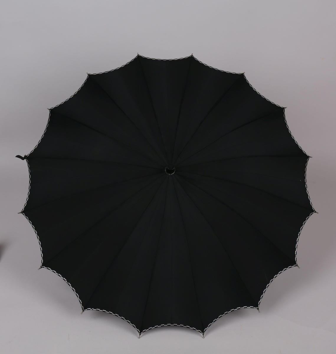 Paraply med 16 metallspilar. Stong i tre. Trekt med stoff i kunstfiber, ensfarga svart med dekor i sølv/svart rutemønster. Handtak i plast med hempe i tekstil til å ta inn på handa. Hempe for lukking av paraplyen.
