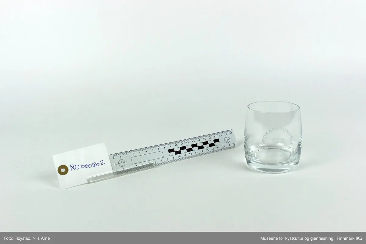 Et rundt brennevinsglass, eller cocktailglass uten stett og trykt tekst på siden av glasset. 

Glasset har en tykk bunn med tynne sider. Øverst står  "NORDKAPPTUNNELEN"  og nederst står "VEIDEKKE". Ordene danner en sirkel og "Gjennomslag 13.5.1998" er trykt inne i sirkelen. Helt nederst står "DYNO".   