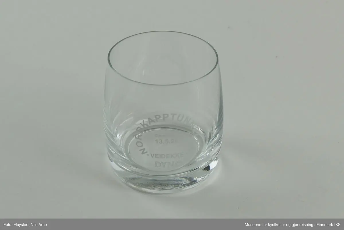 Et rundt brennevinsglass, eller cocktailglass uten stett og trykt tekst på siden av glasset. 

Glasset har en tykk bunn med tynne sider. Øverst står  "NORDKAPPTUNNELEN"  og nederst står "VEIDEKKE". Ordene danner en sirkel og "Gjennomslag 13.5.1998" er trykt inne i sirkelen. Helt nederst står "DYNO".   