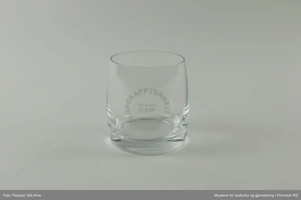 Et rundt brennevinsglass, eller cocktailglass uten stett og trykt tekst på siden av glasset. 

Glasset har en tykk bunn med tynne sider. Øverst står  "NORDKAPPTUNNELEN"  og nederst står "VEIDEKKE". Ordene danner en sirkel og "Gjennomslag 13.5.1998" er trykt inne i sirkelen. Helt nederst står "DYNO".   