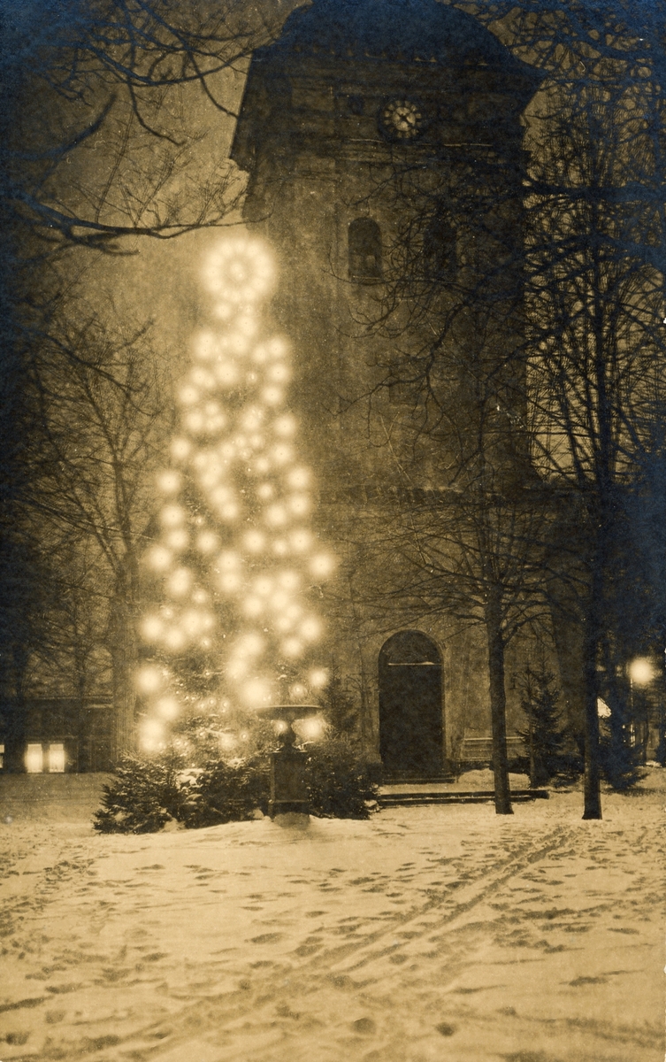 Vykort, "Varberg. Julgranen". Kvällsbild av en tänd julgran i Brunnsparken med kyrktornet i bakgrunden. Marken är täckt av snö. Stämpeln nere till höger försäkrar att kortet är "Äkta fotografi", foto & förlag: Björkström.