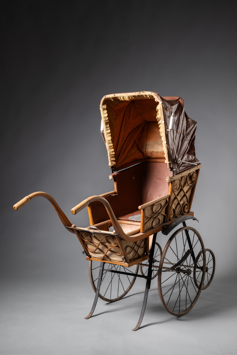 Barnvagn, kombinerad ligg- och sittvagn. Fotlådan går att fälla upp/ner till liggande respektive sittande läge. Stomme av järn bestående av underrede med två stora framhjul och två små bakhjul, samt två stödben (så att vagnen inte tippar fram). Hjulen är av trä med plåtskoning och ekrar av järn. Korg av trä, betsat, dekorerad utvändigt med böjd rotting. Invändigt fodrad med brunt konstläder. Stoppad sits. Två höga, böjda trähandtag. Sufflett av brunt konstläder på metallställning. Suffletten går att fälla upp och ner samt går att ta av. Handtag av vitt porslin. Runt sufflettens ytterkant är ett textilt band med udd fäst. Bandet har ursprungligen varit fäst med nubb, senare fäst med häftstift (de två översta på höger sida i samband med registreringen). Band av textil runt kanten under suffletten.  Sits och fotlådan nötta, skador på suffletten.