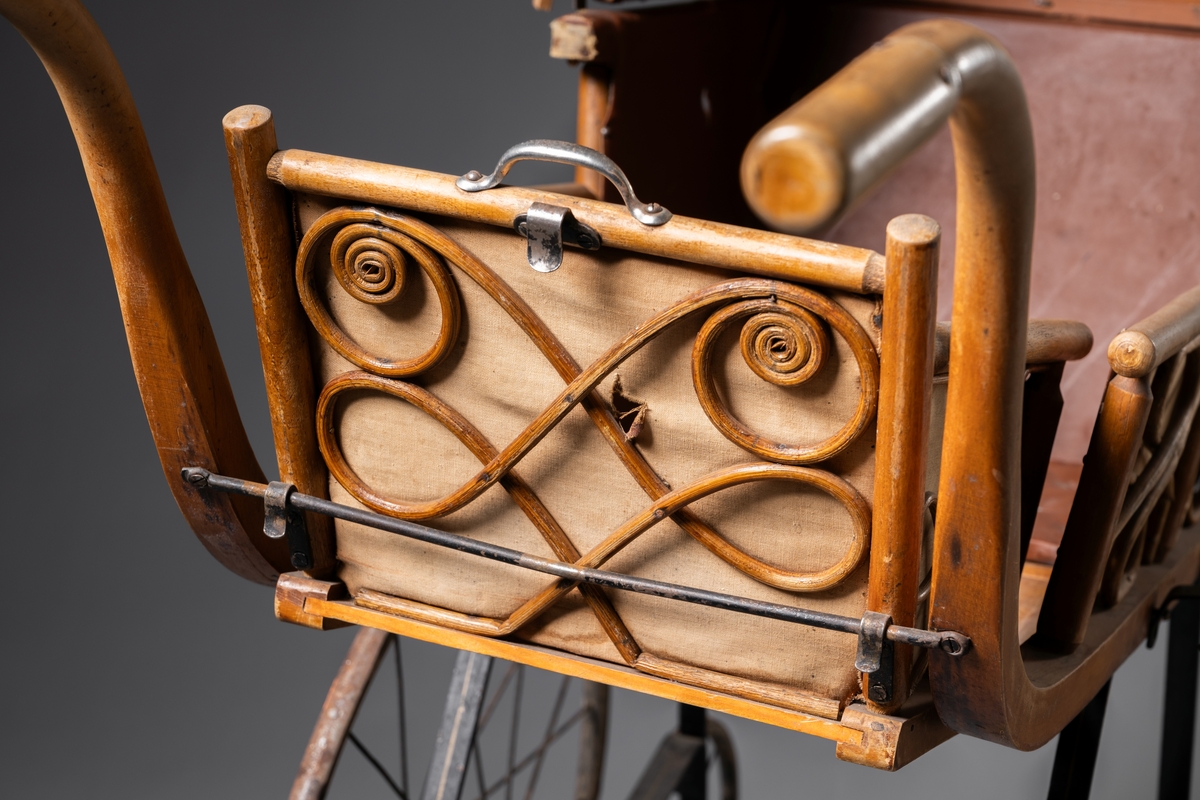 Barnvagn, kombinerad ligg- och sittvagn. Fotlådan går att fälla upp/ner till liggande respektive sittande läge. Stomme av järn bestående av underrede med två stora framhjul och två små bakhjul, samt två stödben (så att vagnen inte tippar fram). Hjulen är av trä med plåtskoning och ekrar av järn. Korg av trä, betsat, dekorerad utvändigt med böjd rotting. Invändigt fodrad med brunt konstläder. Stoppad sits. Två höga, böjda trähandtag. Sufflett av brunt konstläder på metallställning. Suffletten går att fälla upp och ner samt går att ta av. Handtag av vitt porslin. Runt sufflettens ytterkant är ett textilt band med udd fäst. Bandet har ursprungligen varit fäst med nubb, senare fäst med häftstift (de två översta på höger sida i samband med registreringen). Band av textil runt kanten under suffletten.  Sits och fotlådan nötta, skador på suffletten.