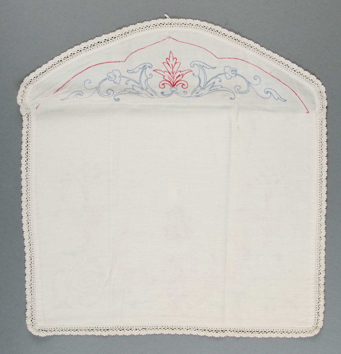 Av vitt bomullslärft med rött och blått broderi i stjälksöm. Kantad med spets. Monogram ES. Knäpps med knapp och ögla.