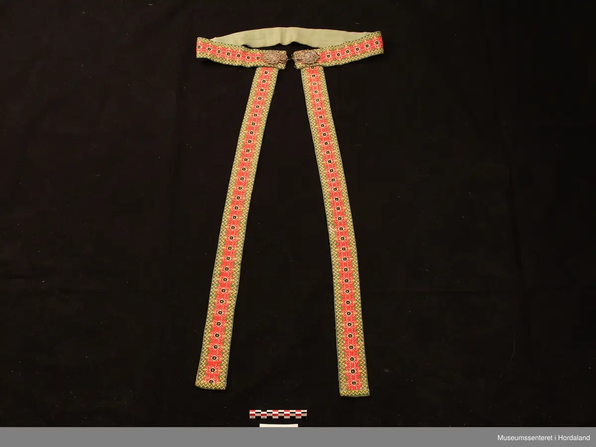 Beltet og fangbanda er vovne i damaskvev. På sidene er det sikk-sakk-border i gult og grønt og midtmønsteret består av  svarte åttebladroser og raude ornament rundt. Lerretsbotnen er kvit. Foret med grønt bomullssateng. Beltet har påsydd ei forgylt sølvspenne i filigransarbeid. Rett under beltespenna er fangbanda festa.