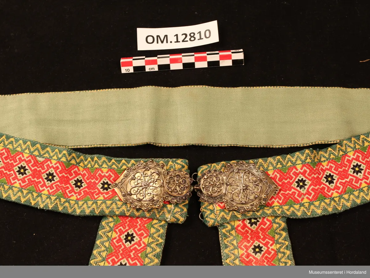 Beltet og fangbanda er vovne i damaskvev. På sidene er det sikk-sakk-border i gult og grønt og midtmønsteret består av  svarte åttebladroser og raude ornament rundt. Lerretsbotnen er kvit. Foret med grønt bomullssateng. Beltet har påsydd ei forgylt sølvspenne i filigransarbeid. Rett under beltespenna er fangbanda festa.