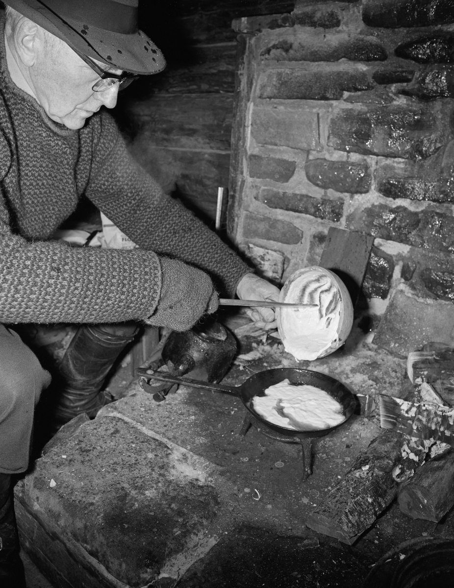 Trygve Aasheim (1893-1975) steker «munk» i damstua fra Kvanstranddammen ved elva Foreninga i Grønavassdraget i Trysil, Hedmark. Fotografiet ble tatt i 1975, fire år etter at koia var flyttet til friluftsmuseet på Norsk Skogbruksmuseum (nå Norsk Skogmuseum) i Elverum (jfr. SJF-B.0026). Vi ser Aasheim, stående bøyd framfor peisen, der han helte munkrøre fra en auskopp ned i ei stekepanne. Om munken skriver Geir Hovensjø i Norsk Skogbruksmuseums årbok nr. 7, der fotografier av Aasheim i Kvannstranddamkoia var brukt som illustrasjoner: 

«Munk var flere steder hovedretten i kostholdet. (Trysil, Åmot, Hedmark). Munkerøra besto av hvetemel utrørt i vatn og tilsatt salt. Munken ble stekt i fleskefett eller smult til ei tjukk pannekake. Enkelte brukte natron eller eggepulver til hevelse. Røra ble slått i en ring i panna så det ble en åpning i midten for fettet, slik at munken nærmest fløt i panna.

Om munkens betydning forteller Birger Nysæther [1918-2001] fra Åmot: «Munk var obligatorisk midt på dagen – og hver dag hele vintersesongen igjennom. Ingen ble lei av denne kosten, selv om det foregikk gjennom alle år. En kunne variere noe med det en drakk til – melk, kaffe eller en kunne også spise «typpærsoll» ved siden av. På munken brukte en aldri syltetøy, bare sukker. Munkspade var noe en laget allerede første dagen i koia.»

Aasheim var museets kontaktmann i Trysil. Hans engasjement og kontaktnett i Trysil var viktige forutsetninger for at dette husværet ble flyttet til museet og bevart der. Aasheim bidro også, sammen med sambygdingen Jon Galaasen (1922-2002), med praktisk både da koia skulle demonteres i Trysil og da den ble gjenoppført på ei tomt i nærheten av dammen nederst i  fløtingsåa som ble opparbeidet på sørsida av flomvollen mot Prestfossen, i den nordre delen av friluftsmuseet på Prestøya. Damanlegget ble ferdig i 1974. Aasheim ivret også for formidling av damfløternes hverdagsliv og skaffet cirka 40 gjenstander som skulle gi publikum en illusjon av at koia var bebodd.