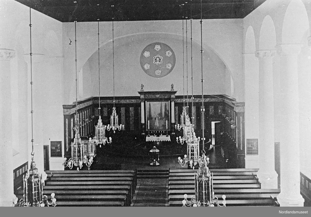 Kirkeinteriør, Narvik kirke, sett fra galleriet, lysekroner. Åtte lysekroner, fire på hver side. Døpefont midt foran alterringen. Veggene rundt alteret er dekorert. Til venstre foran, prekestolen med utsmykninger.