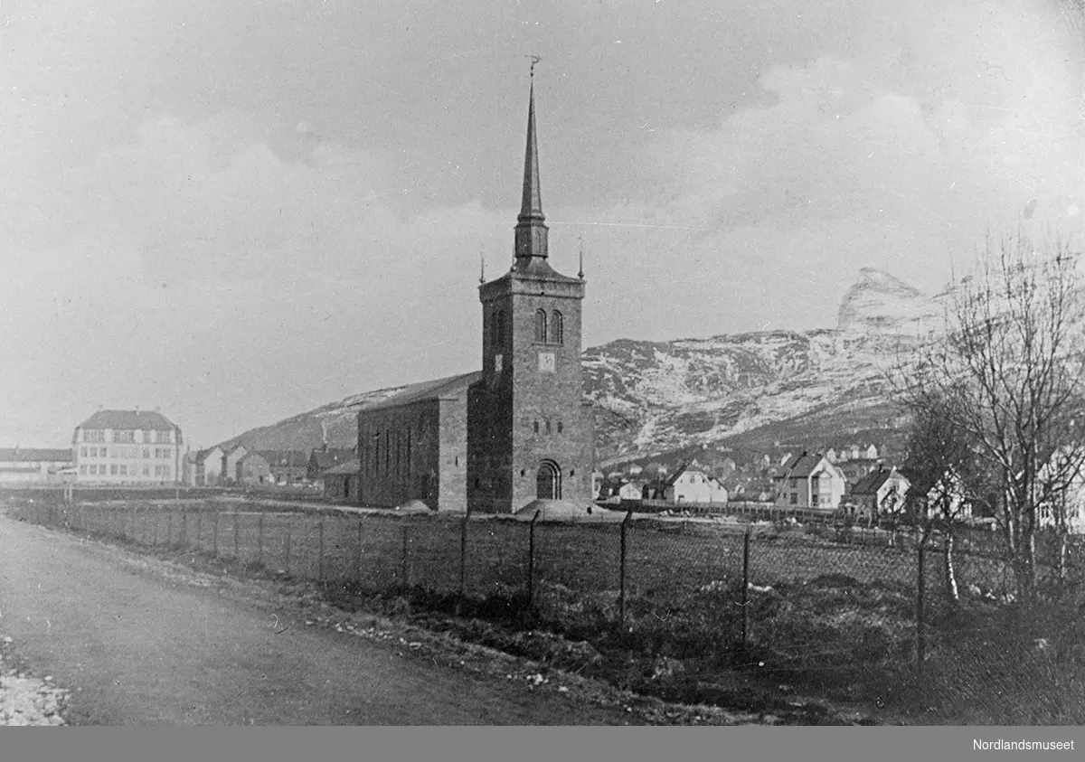 Narvik kirke, sett fra Framnessiden, foran et gjerde. Kirken er bygd i stein med spir og værhane. Gaten til høyre er Kirkegaten. I bakgrunnen ses bydelene Frydenlund og Oscarsborg. Fjellet i bakgrunnen er Fagernesfjellet.