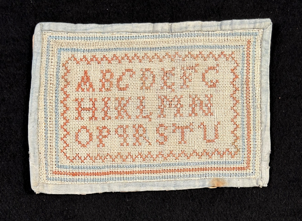 Märkduk av tuskaftat linne, sydd med korssöm. Alfabetet i mitten , bård runt om i rosa och ljusblått. Kantad med ljusblått tuskaftad bomull. Uppfodrad och fastsydd på papper.