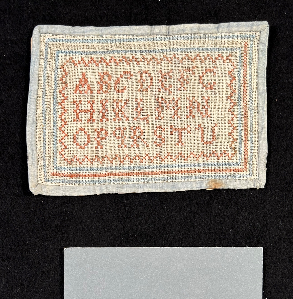 Märkduk av tuskaftat linne, sydd med korssöm. Alfabetet i mitten , bård runt om i rosa och ljusblått. Kantad med ljusblått tuskaftad bomull. Uppfodrad och fastsydd på papper.