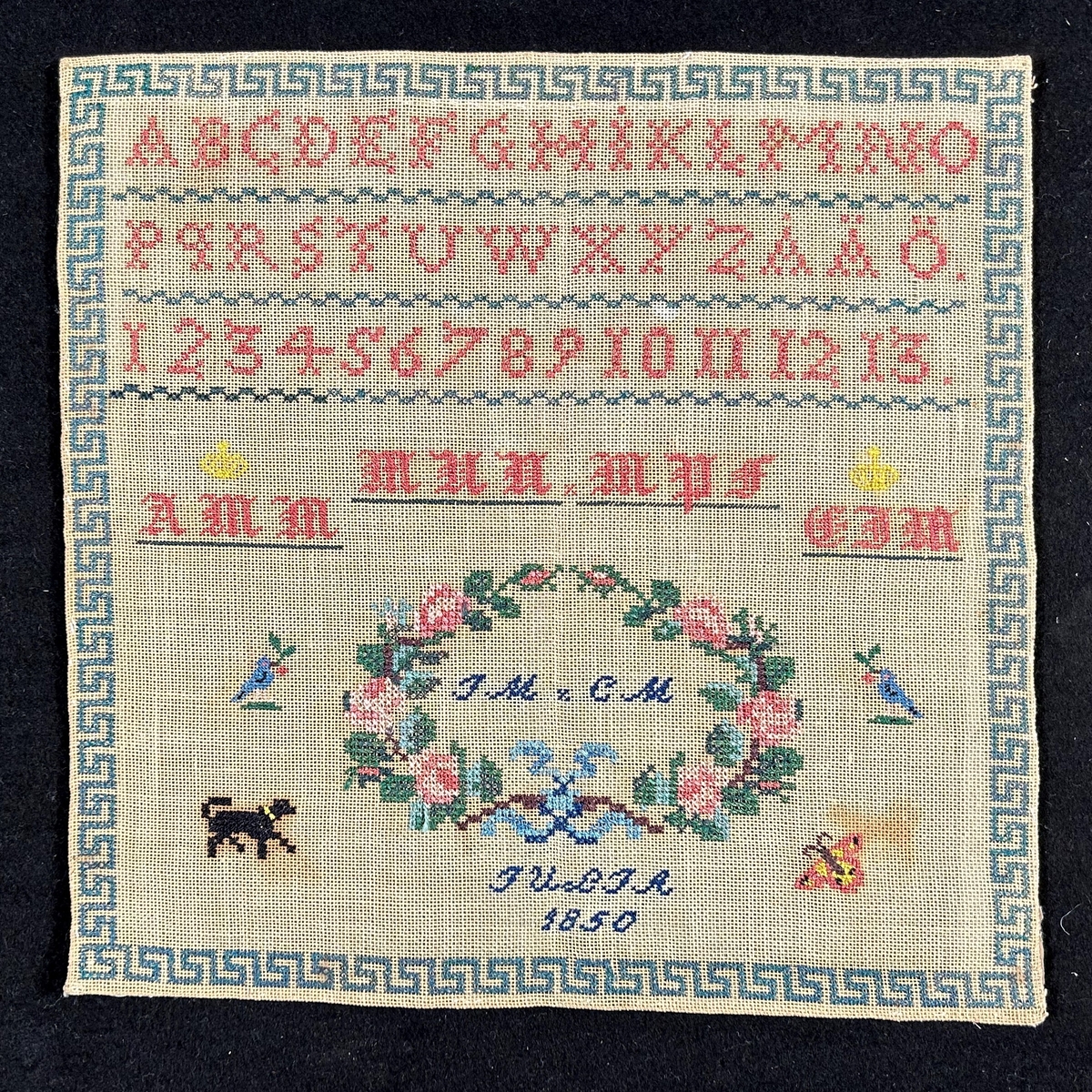 Märkduk med botten av naturfärgad linne. Broderi av silke i färgerna rött, grönt, blått, svart och gult. Längst upp alfabetet samt en sifferrad 1-13, totalt tre rader. Monogram i frakturstil; (krönt)AMM, MNN, MPF, (krönt)CIM. Inom blomsterkrans: IM, CM. Längst ned JULIA 1850. Runt om en bård av meandermönster.