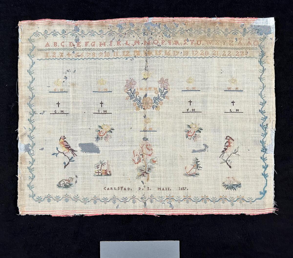 Märkduk på botten av ofärgad appreterad ylle. Korsstygngsbroderi i pastellfärger; blått, rosa, och brunt. Bård rut om. Upptill två rader med alfabetet (romersk kapitälskrift), samt siffror. Symmetrisk komposition. en rad med krönta monogram; GH, IH, IHCMH, CH, WH. En rad med korsprydda monogram; GH, IH, FH, CH. Krönt monogram CH i mitten. Längst ner: CARLSTAD. D. 3. MAII. 1839. Spridda blom- och figurmotiv.