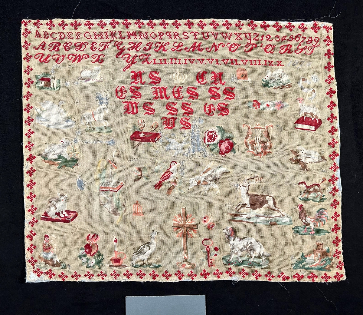 Märkduk med broderi på beige stramalj. Färger i rött, grönt, brunt och vitt. Tre bårder upptill med alfabetet i olika stilar, siffror (även romerska). Under en vit broderad krona, monogram i frakturstil. Strödda motiv över hela duken: blommor, djur, böcker, lyra, nyckel, ljus, med flera symboler. Längst ned i mitten ett kors.