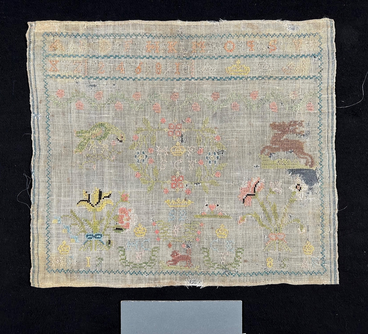 Märkduk av naturfärgat ylle, korssömsbroderier, kantbård runt om. Symmetrisk komposition. Överst två rader med bokstäver i alfabetisk ortdning, sifferrad samt fyra olika typer av kronor. Djur och blommotiv i färgerna grönt, rosa, brunt, grått, gult och blått.