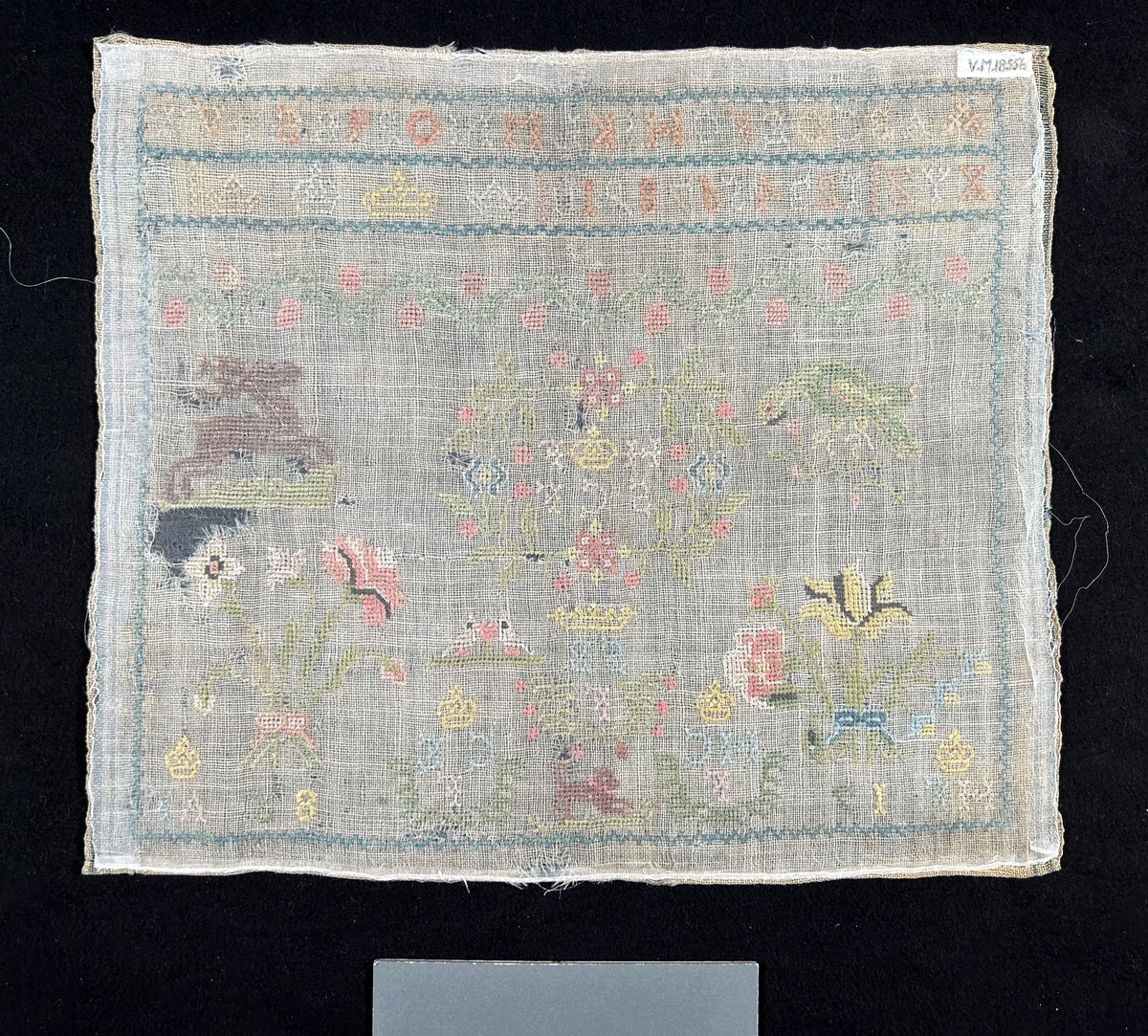 Märkduk av naturfärgat ylle, korssömsbroderier, kantbård runt om. Symmetrisk komposition. Överst två rader med bokstäver i alfabetisk ortdning, sifferrad samt fyra olika typer av kronor. Djur och blommotiv i färgerna grönt, rosa, brunt, grått, gult och blått.