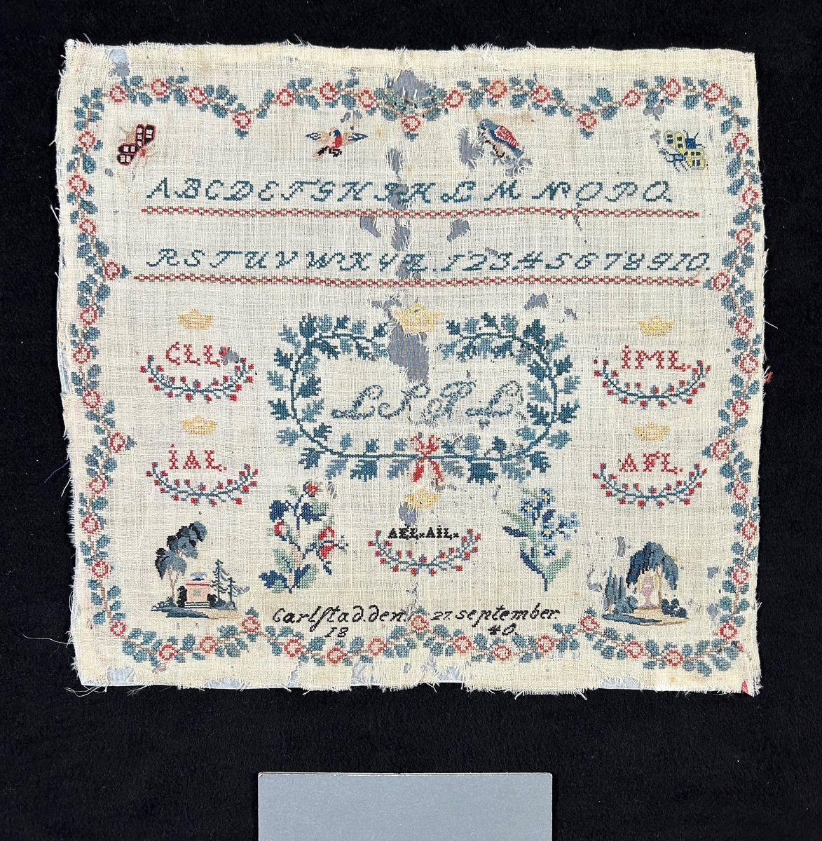 Märkduk med blomsterranka runt kanten. Två rader längst upp med alfabetet i engelsk skrivstil och siffror. Symmetrisk komposition. I mitten krönt inramat monogram; LSPL. På vänster sida två krönta monogram; CLL och IAL, på höger sida; IML och AFL. Under det stora monogrammet i mittet, två stycken monogram under en krona; AEL och AIL. Längst ned daterad: Carlstad den 27 september 1840. Övrigt spridda blommotiv, fåglar och fjärilar, träd och vyer.