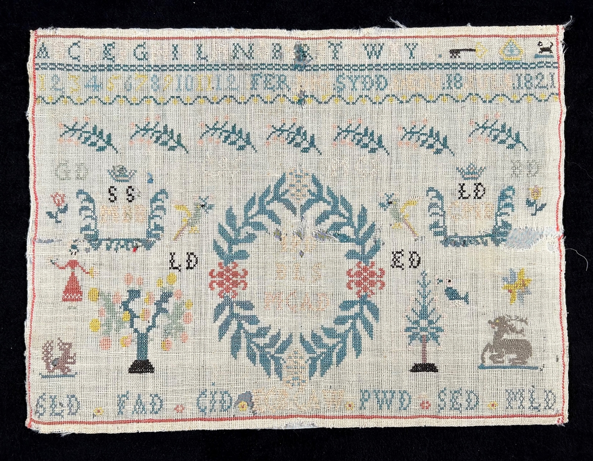 Märkduk i symmetrisk komposition. Överst två rader med alfabetet och siffror. På den undre raden står "Ferdigsydd den 18 juli 1821." I mitten en oval krans med monogram; JPD, BLLS, MCAD, på båda sior två par krönta monogram SS, MBB LD, CMB. Längst ned en rad med olika monogram; SLD, FAD, CJD, ECB, CAW, PWD, SED, MLD. För övrigt växt- och djurmotiv. Monterad på papper.
